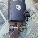 Černý obdélníkový box s nápisem SnareBox a logem MEINL. Box je připevněn k kovovému pedálu s šedým plastovým stupňem, na kterém je logo Remo. Povrch…
