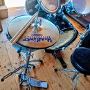 Fotografie zobrazuje část bicí sady na dřevěné podlaze. Na činelym talíři je modrý nápis Headliner a PERCUSSION. Součástí je hi-hat stojan s pedálem,…