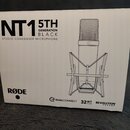 Fotografie znázorňuje bílé originální balení mikrofonu. Na krabici je černý nápis NT1 5TH GENERATION BLACK STUDIO CONDENSER MICROPHONE a logo RØDE. V…