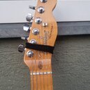 Fotografie zachycuje hlavu kytary z jasného dřeva s šesti chromovanými ladičkami. Na povrchu je vidět nápis "Fender" a modelové označení "TELECASTER"…