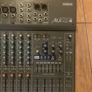 Šedý mixovací pult značky YAMAHA s označením Mx12/4 MIXING CONSOLE. Zařízení obsahuje řadu vstupů, posuvníků a ovládacích knoflíků s popisy jako GAIN…