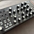 Černý semi-modulární analogový syntetizátor s dřevěnými bočními panely. Na předním panelu je bílý nápis SUBHARMONICON | SEMI-MODULAR ANALOG POLYRHYTH…
