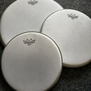 Tři bílé kruhové bubnové blány s kovovými obručemi. Každá blána má v horní části černé logo REMO a pod ním nápis EMPEROR® WHITE COATED MADE IN U.S.A.…