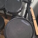 Elektronická bicí souprava v černé barvě. Na povrchu padů jsou viditelná loga CARLSBRO. K soupravě patří dřevěné paličky umístěné v držáku na stojanu…
