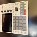 Bílá MPC produkční stanice s šedými pady a černým displejem. Na horním levém rohu je červené logo AKAI professional a vlevo u displeje nápis MPC ONE…