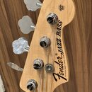 Fotografie zachycuje hlavu basové kytary z přírodního světlého dřeva s четыřmi chromovanými ladičkami. Na povrchu je černý nápis Fender® JAZZ BASS a…