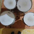 Fotografie zachycuje čtyři bílé bubny bicí s kovovými obručemi a jeden činely. Na dvou z bubnů je viditelné logo REMO, na jednom je nápis EVANS. Povr…