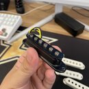 Černý humbucker snímač s šesti kovovými póly, který drží ruka. Snímač má připojené žluté a černé vodiče. V pozadí jsou vidět tři další bílé snímače.…