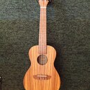Dřevěný nástroj typu ukulele s přirozenou barvou a výraznou sletovanou strukturou dřeva. Na hlavě je viditelné logo ve tvaru stylizovaného písmena „C…