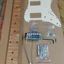 Sada komponentů pro elektrickou kytaru položená na hnědé kartonové ploše. Obsahuje dřevěný krk s hnědým lakem, bílý plastový pickguard a bílou krytku…