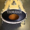Světle žlutý basový buben s černým předním koženem. Na koženě je bílý nápis YAMAHA a uprostřed je vyříznutý otvor. Nástroj má chromované lugy a nožič…