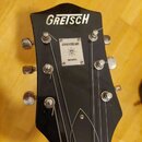 Fotografie zachycuje hlavu černé kytary s bílým nápisem GRETSCH. Uprostřed je umístěn stříbrný čtvercový štítek s nápisy ANNIVERSARY a MODEL, mezi kt…