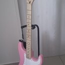 Růžová elektrická kytara s bílým pickguardem a světle hnědým hnedkem. Na hlavě je viditelné logo Squier a nápis SERIES STRATOCASTER. Nástroj je umíst…