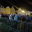 Fotografie nezobrazuje žádný hudební nástroj ani příslušenství. Je na ní vidět skupina lidí za kovovými zábranami na nočním městském náměstí s řadou…