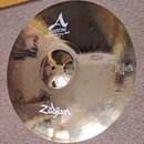 Zlatý kovový činel s viditelným kování povrchu. Na horní části je bílý nápis A CUSTOM 20th Anniversary Ride MEDIUM THIN 22" 55 cm a ve spodní části b…