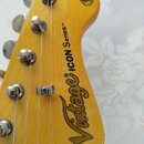 Detail hlavy elektrické kytary z jasnozlatého dřeva s lakovaným povrchem. Na dřevě je černý nápis Vintage® ICON Series™ a v horní části kulatý znak s…