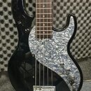 Černá elektrická basová kytara s hnedým hmatníkem. Nástroj má světle šedý pickguard s perleťovým vzorem a čtyři ladicí knoflíky. Na těle nejsou vidit…