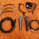Sada vybavení složená ze dvou dynamických mikrofonů, kabelů a napájecího adaptéru. Na mikrofonech jsou viditelné nápisy "Sennheiser" a "Cobalt 810".…