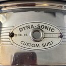 Detail kovového, chromovaného povrchu nástroje s oválným štítkem. Na štítku je nápis DYNA-SONIC a CUSTOM BUILT, vprostřed je otvor a vlevo text SERIA…