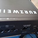 Černý elektronický hudební přístroj s bílým nápisem KURZWEIL na horní straně. Na zadním panelu jsou konektory s popisky MIDI OUT, MIDI THRU a MIDI IN…