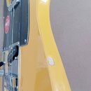 Detail žluté elektrické kytary s černým pickguardem a chromovanými komponenty. Na boku těla nástroje je viditelný výrazný odřap s chybějícím lakem a…