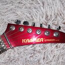 Červená hlava elektrické kytary s šesti kovovými ladičkami. Na povrchu je nápis KRAMER TM STRIKER 100 ST. Lak je v několika místech poškrábaný a vyka…