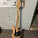 Elektrická basová kytara s přírodním dřevěným povrchem těla a hmatníku. Nástroj má černý pickguard, dva snímače a čtyři ladicí kolíky na hlavě. Na hl…