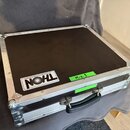 Černý transportní kufr s hliníkovými hranami a kovovými rohy. Na přední straně je bílá náleplka s nápisem THON a menším textem "Professional flight e…