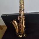 Zlatý saxofon umístěný v černém otevřeném kufru. V popředí leží samostatně vyjmutý munzpun s trškou. Na povrchu nástroje nejsou viditelná žádná loga…