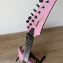 Růžová elektrická kytara s šesti strunami, umístěná na černém stojanu. Na hlavě nástroje je zlatý nápis Ibanez. Povrch kytary vypadá hladce a bez vid…
