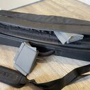 Černý obal na hudební nástroj s několika zipy a přivázaným popruhem. Na černé části popruhu je šedý nápis D'Addario. Obal je mírně otevřený a uvnitř…