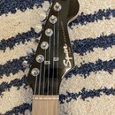 Fotografie zachycuje hlavu elektrické kytary v černé barvě. Na povrchu je bílý nápis Squier STRATOCASTER® a pod ním menší logo Fender. Nástroj má šes…