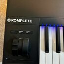 Černý klaviatura kontrolér s bílými a černými klávesami. Na horní části je bílý nápis KOMPLETE s doprovodným logem. Pod ním se nacházejí tři tlačítka…