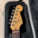 Hlava elektrické kytary z přírodního dřeva v otevřeném hliníkovém kufru s černým polstrováním. Na hlavě je černé logo Fender a nápis STRATOCASTER ora…