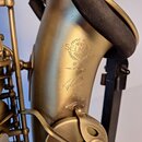Detail saxofonu v matné zlaté barvě. Na korunu je vyryto logo s nápisem HENRY SELMER PARIS, symbol ® a text MADE IN FRANCE. Pod nimi se nachází ruční…