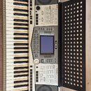 Bílo-šedý digitální klavír s černým notovým stojanem. Na přístroji je viditelné logo YAMAHA a nápis PSR-2000. Instrument obsahuje klávesnici, displej…