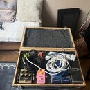 Otevřený dřevěný kufr s šedým vnitřním polstrováním, který obsahuje kytarový pedalboard. Jsou zde vidět efekty značek Boss a další pedály, z nichž je…