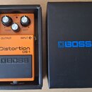 Oranžový efekt pedál v černé krabici. Na pedálu jsou nápisy Distortion DS-1 a logo BOSS, dále označení ovládacích prvků TONE, LEVEL a DIST s nápisem…