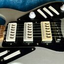 Detail elektrické kytary v stříbrné barvě s černým pickguardem. Nástroj má tři snímače typu single-coil v bílé a krémové barvě. Na těle jsou vidět bí…