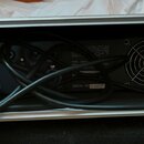 Zadní strana černého elektronického zařízení umístěná v hliníkovém transportním kufru. Na zadním panelu je vidět ventilátor s mřížkou a nápis SERIAL…