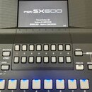 Černý digitální klavír značky YAMAHA s displejem, na kterém je nápis PSR-SX600. Na obrazovce jsou dále viditelné texty Program Version 1.03, Hardware…