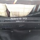 Zadní strana černého elektronického klavíru s nápisem Roland E-70. Jsou vidět napájecí vstup, několik zapuštěných konektorů a varovné štítky s textem…