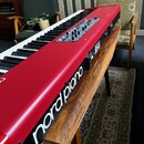 Červený digitální klavír s bílými a černými klávesami, umístěný na dřevěném stole. Na boku je bílý nápis "nord piano". Horní panel obsahuje ovládací…