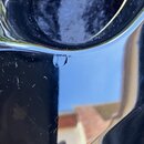 Detailní záběr na černý, vysoce lesklý povrch hudebního nástroje. V horní části je vidět zaoblená hrana s odrazem okolí. Na povrchu jsou patrné drobn…