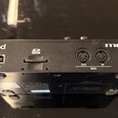 Zadní strana černého elektronického přístroje s nápisy Roland a MC-101. Jsou zde vidět přepínač POWER (ON/OFF), USB port, slot pro SD kartu s označen…
