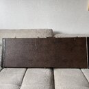 Hnědý obal na hudební nástroj v obdélníkovém tvaru s bílým lemováním po okrajích. Povrch vykazuje známky opotřebení v podobě škrábanců a odřenin. Na…