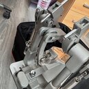 Fotografie zachycuje detail šedého kovového mechanismu bicího pedálu. Konstrukce obsahuje pružiny, klouby a nastavitelné šrouby. Na povrchu nejsou vi…