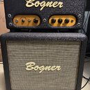 Kytarový zesilovač a reproduktorové boxy značky Bogner. Horní jednotka má zlatý ovládací panel s nápisy BARCELONA, volume, gain, bass a treble. Předn…