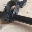 Černý kovový držák s dvěma utahovacími knoflíky, který je upevněn na černé kovové tyči. Na boku držáku je vyražen nečitelný nápis. Celý povrch je mat…