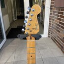 Hlava a hриф elektrické kytary z jasného dřeva umístěné na černém stojanu s nápisem HERCULES. Na hlavě je černé logo Fender a nápis STRATOCASTER. Pod…